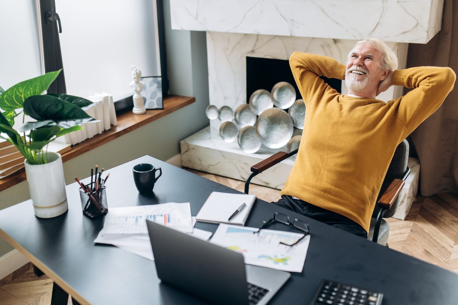 A gray-haired smiling senior man leaned on an armchair holding his hands behind his head while resting after work in a laptop